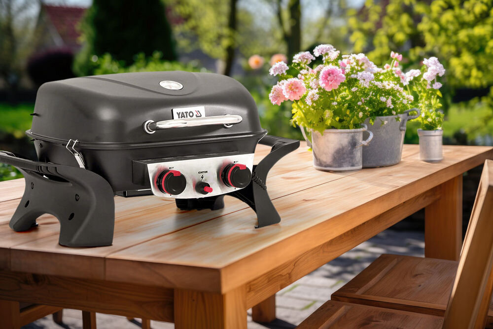 Grill gazowy YATO YG-20005 Yake Czarny 5 kW 47 x 37 cm, w tle taras i stół ogrodowy, na pierwszym planie grill gazowy na stole