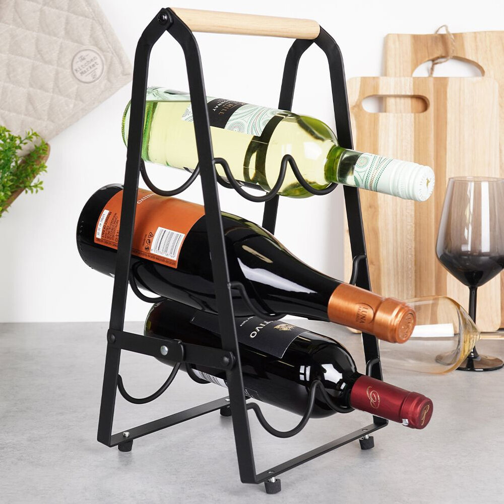 Czarny metalowy stojak na wino z drewnianym uchwytem, mieszczący trzy butelki wina. W tle widoczne są deski do krojenia, kieliszek oraz roślina.