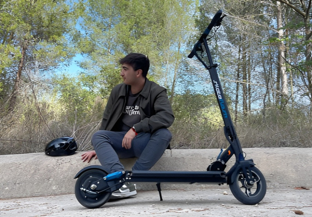 Hulajnoga elektryczna SKATEFLASH Echo 40km 350W 8 cali Czarny droga park las hulajnoga stoi na drodze oparta na stopce w tle na murku siedzi osoba patrzy w bok widoczna od przodu pod skosem składana stopka wytrzymała nóżka w centralnym punkcie ciężkości