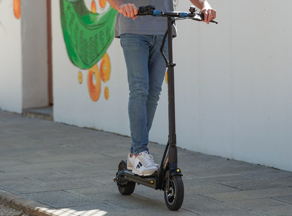 Hulajnoga elektryczna SKATEFLASH Echo 40km 350W 8 cali Czarny miasto chodnik osoba jedzie na hulajnodze po chodniku w tle budynek amortyzator tłumi drgania i wstrząsy płynna i bardziej stabilna jazda lepsza przyczepność kół do podłoża