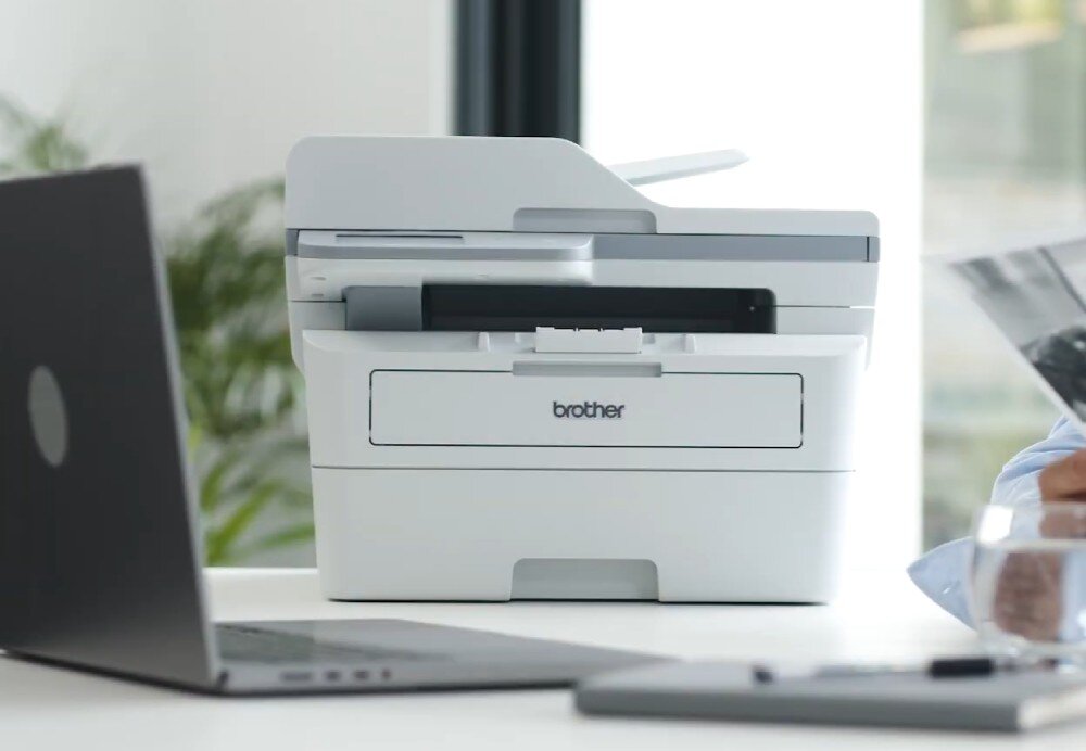 Urządzenie wielofunkcyjne BROTHER MFC-L2922DW urządzenie stojące na stole wraz z laptopem i notatnikiem głośność podajnik na papier