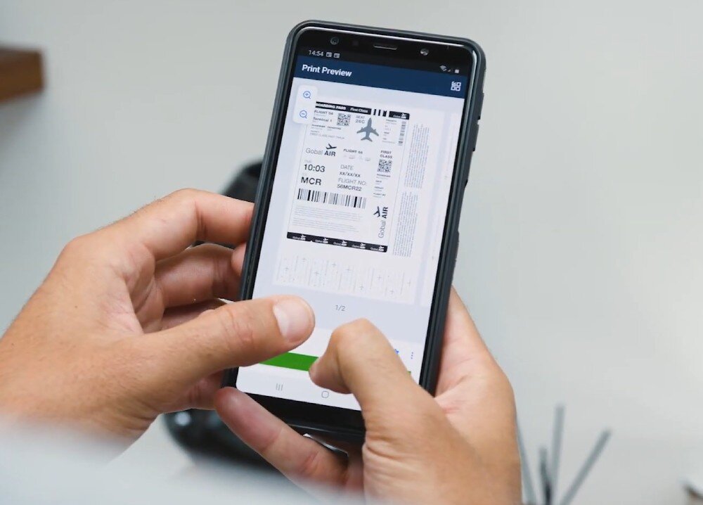Urządzenie wielofunkcyjne BROTHER MFC-L2802DW zbliżenie na osobę trzymającą smartfon z widocznym ekranem, na którym widać aplikację z przygotowaniem biletu lotniczego do druku łączność Wi-Fi Ethernet USB aplikacja
