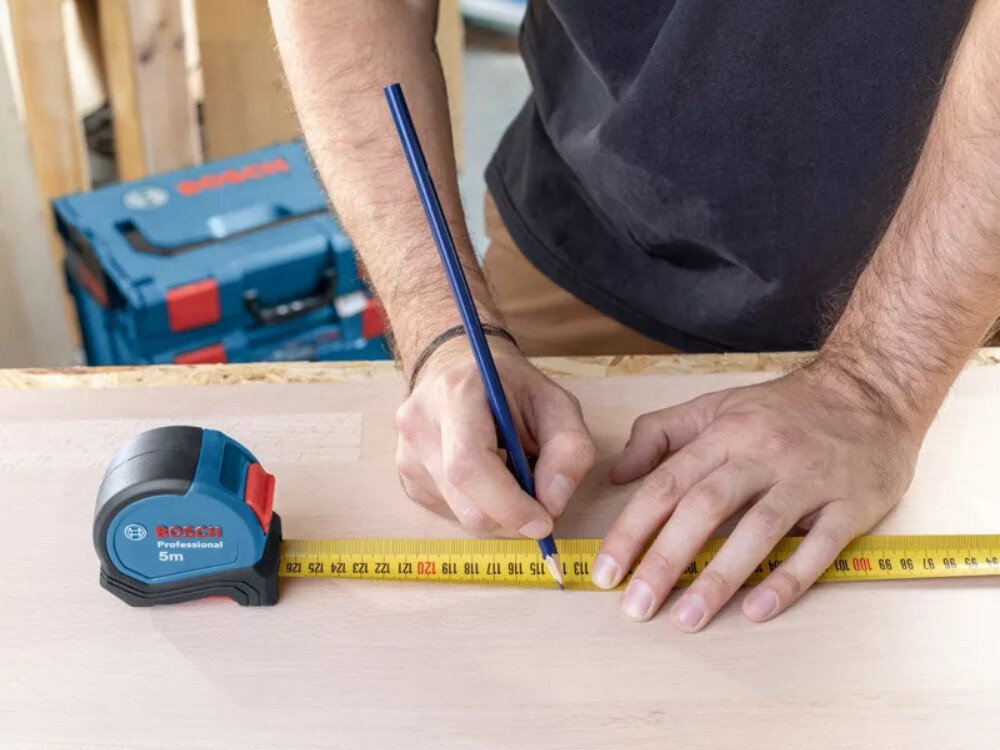 Miara zwijana BOSCH Professional 1 600 A01 6BH (5 m), miara, Bosch, pomiar, budowa, osoba odmierza dystans miarą na desce na stole stolarskim