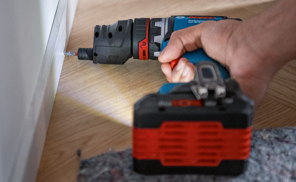 Wiertarko-wkrętarka BOSCH GSR 18V-90 FC Professional (06019K6200) drewniana podłoga osoba przykręca śrubkę do listwy przyściennej widoczne oświetlenie wbudowane oświetlenie LED doświetla miejsce pracy pozwala szybciej i dokładniej ustawić nasadkę na śrubie zmniejszając ryzyko poślizgu i uszkodzenia elementów
