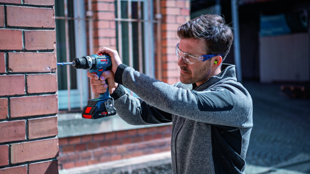 Wiertarko-wkrętarka BOSCH GSR 18V-90 FC Professional (06019K6202) dom ściana z cegły osoba wierci otwór w ścianie w tle okna wykonywanie szerokiego zakresu prac montażowych i budowlanych wymienne uchwyty jedno narzędzie zastępuje kilka urządzeń