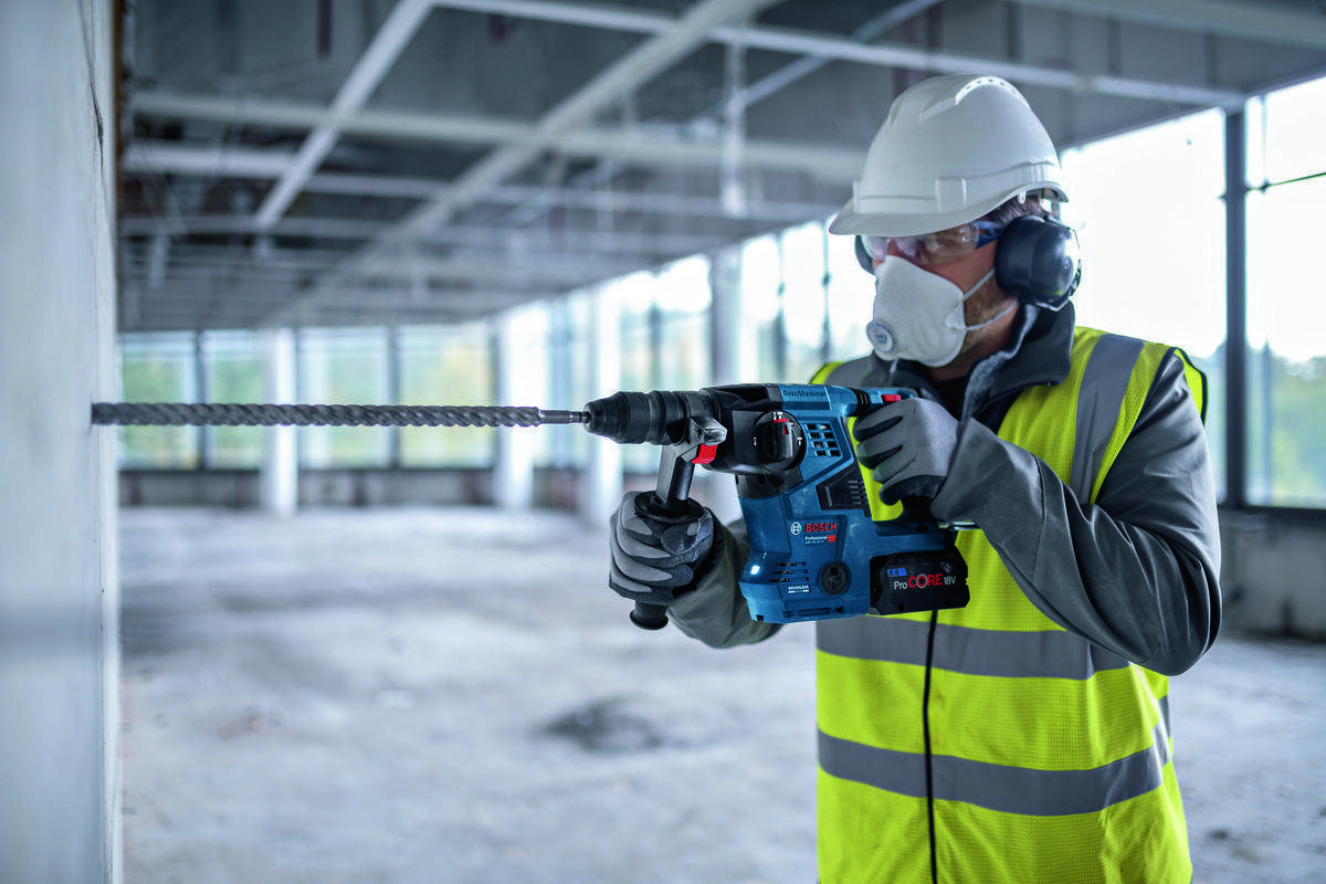 BOSCH GBH 18V-28 C BOSCH GBH 18V-28 C beton, cegła czy kamień wykonywać otwory dłutować fragmenty powierzchni - Mężczyzna w okularach ochronnych wierci otwór w ścianie za pomocą akumulatorowego młota udarowo-obrotowego