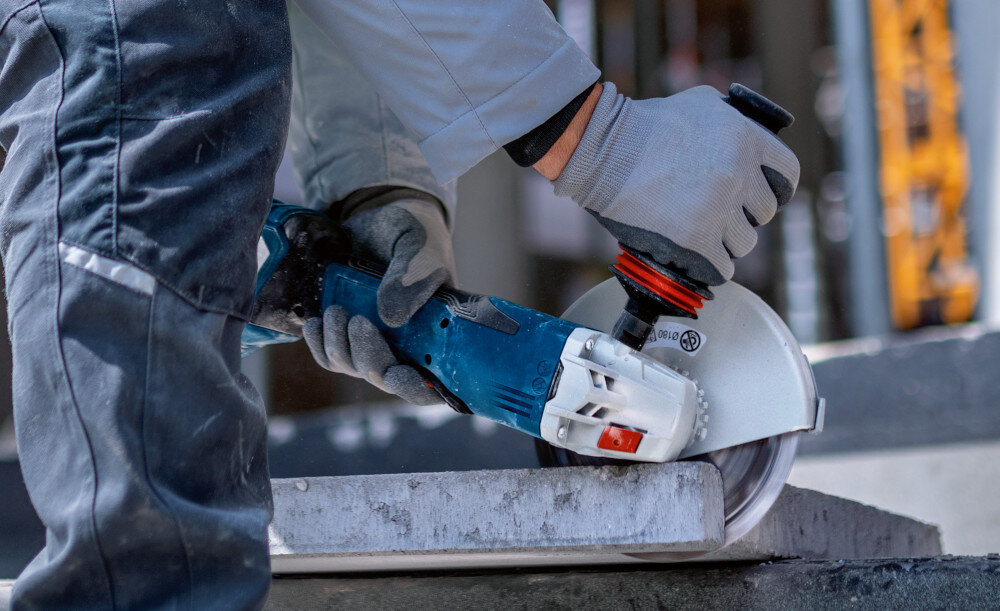 Szlifierka kątowa BOSCH Professional GWS 18V-180 P solo 0 601 9H6 L01 osoba trzyma szlifierkę w dłoniach tnie beton bezpieczeństwo użytkowania wbudowany system zapobiegania przypadkowemu włączeniu po zaniku prądu ochrona przed przeciążeniem wyłącznik przeciążeniowy ochrona narzędzia przed przegrzaniem