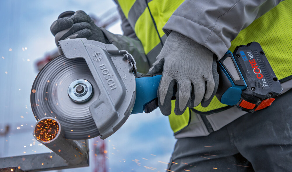 Szlifierka kątowa BOSCH Professional GWS 18V-180 P solo 0 601 9H6 L01 osoba używa szlifierki do cięcia metalowej rury przycisk blokady wrzeciona M14 szybka zmiana tarczy nie wymaga zastosowania jakichkolwiek narzędzi współpracuje z tarczą o średnicy 180 mm w zestawie dwie tarcze tnące Expert for Inox i tarcza diamentowa