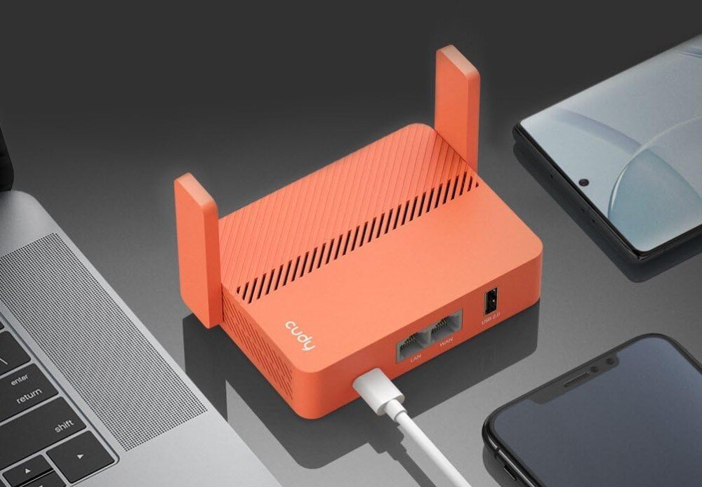 Router CUDY TR1200 2.4 / 5 GHz Mały pomarańczowy router Wi-Fi z dwiema składanymi antenami stoi na biurku obok laptopa i dwóch smartfonów, opis