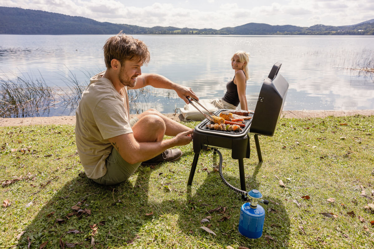 Grill gazowy CAMPINGAZ Tour and Grill 2.4 kW 47 x 25.5 cm, gotowanie, smażenie, ogród, podróż, mężczyzna obsługuje grill, w tle kobieta korzystająca z wypocznku na łonie natury