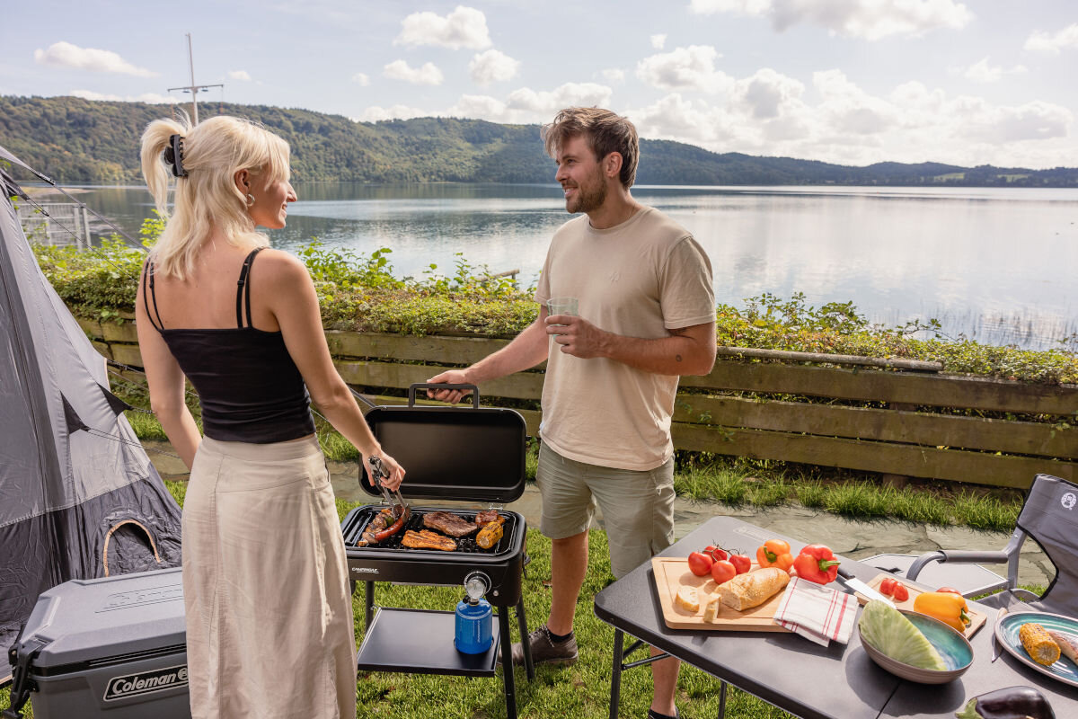 Grill gazowy CAMPINGAZ Tour and Grill 2.4 kW 47 x 25.5 cm, powierzchnia, wymiary, emaliowana, stabilne, w tle jezioro, para rozmawia przy grillu