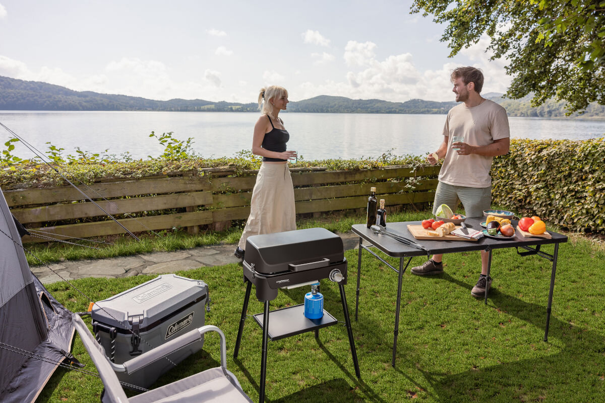 Grill gazowy CAMPINGAZ Tour and Grill 2.4 kW 47 x 25.5 cm, zamontować, nogi, półka, pozycja, w tle jezioro, para rozmawia, na pierwszym planie grill z zamkniętą pokrywą