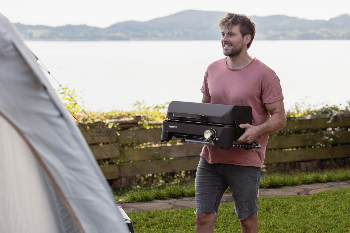 Grill gazowy CAMPINGAZ Tour and Grill 2.4 kW 47 x 25.5 cm, zintergowane, uchwyty, przenoszenie, wymiary, mężczyzna przenosi grilla w rękach, w tle jezioro