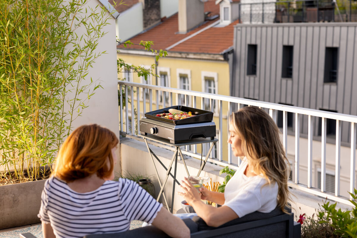 Grill elektryczny CAMPINGAZ Plancha Electric, wymiary, stabilne, waga, wygoda, bezpieczeństwo, 2 kobiety siedzą a sofie i rozmawiają na balkonie, w tle grill na stojaku