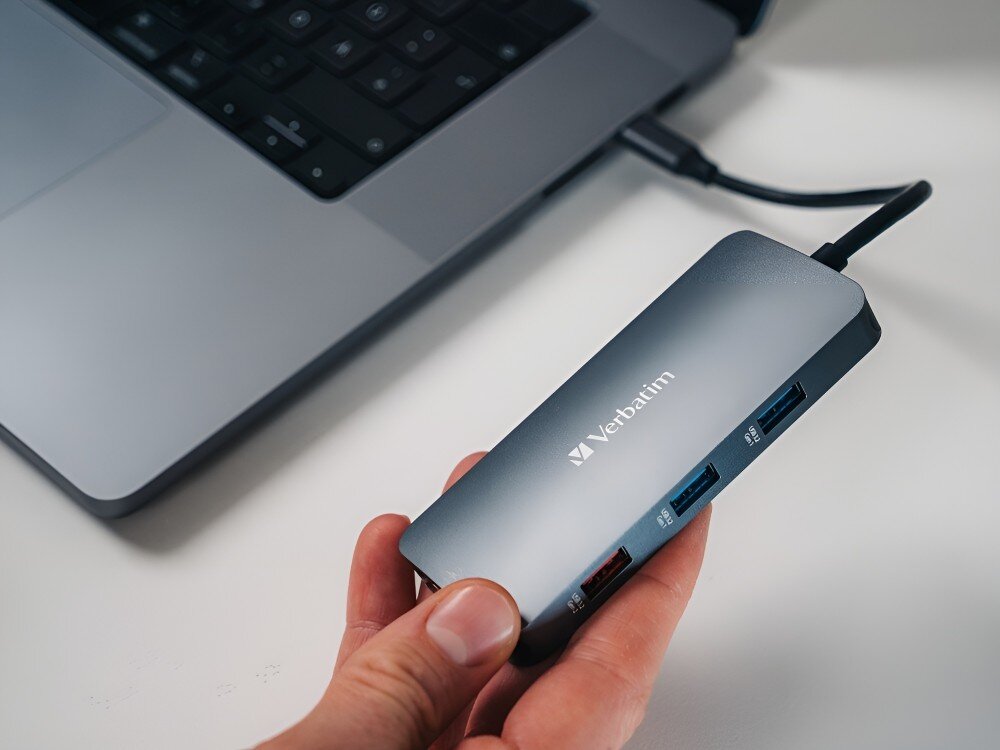 Hub Verbatim 32152 Osoba trzyma adapter z kilkoma portami USB, podłączony do laptopa leżącego na biurku, 
            hub gadżet technologia adapter laptop komputer