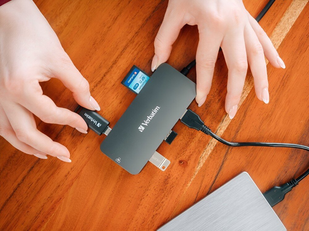 Hub Verbatim 32152 Kobieta wkłada pendrive do adaptera leżącego na drewnianym biurku, obok karta SD i inne podłączone przewody, 
            hub prędkość 10 gb/s gen2 power delivery pd