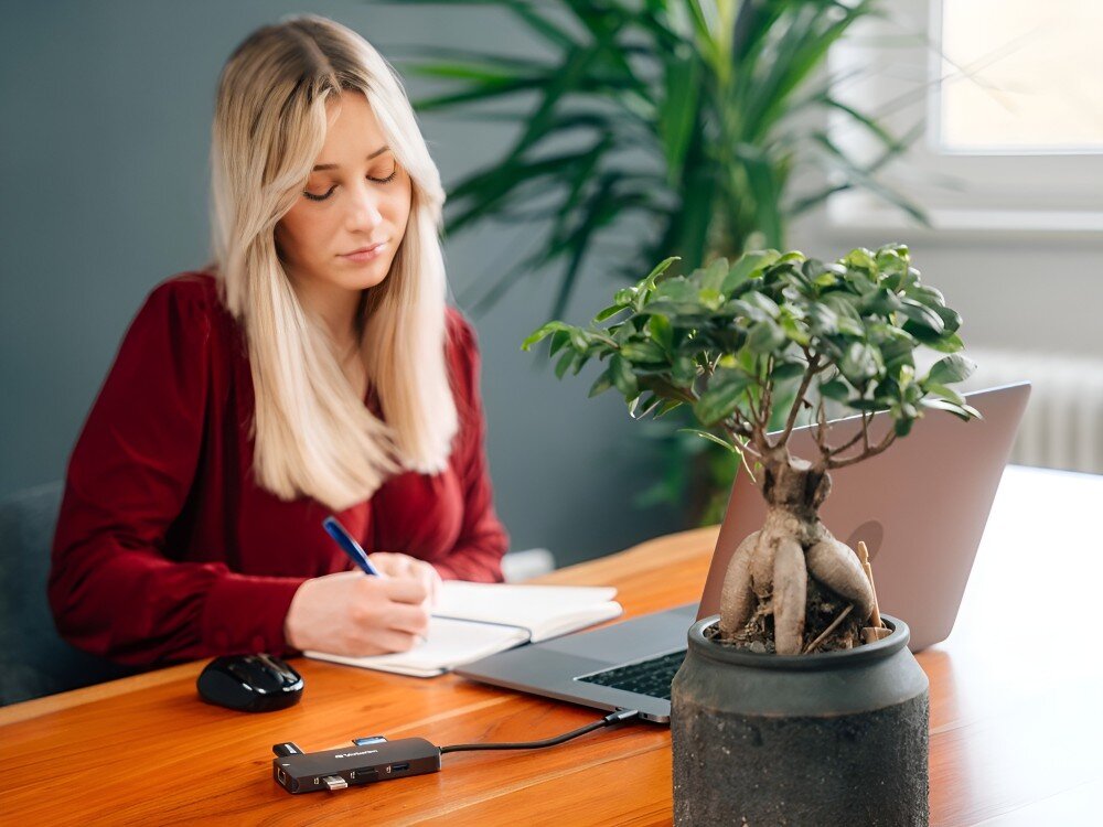 Hub Verbatim 32152 Kobieta w czerwonej bluzce siedzi przy biurku i pisze w notesie, obok otwartego laptopa i podłączonego adaptera; na pierwszym planie widać doniczkę z rośliną,
            hub sterowniki usbc kompatybilność windows macos chrome os linux android