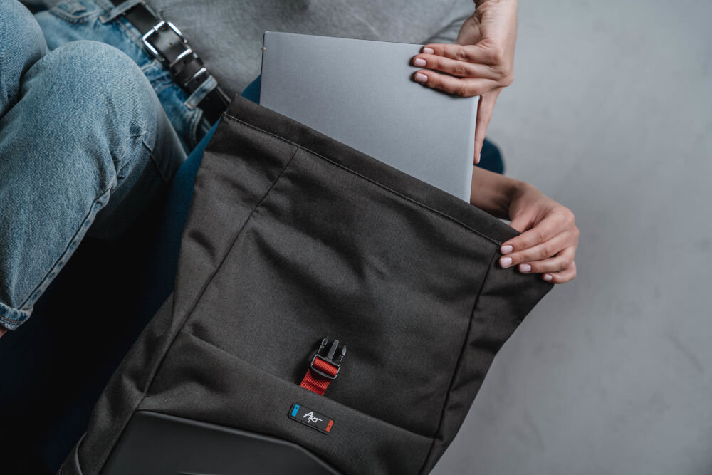 Plecak na laptopa ART BP-9367 15.6 cali prezentacja funkcji roll-top kobieta wkładająca laptopa do plecaka w pokoju siedząca na niebieskiej kanapie powiększ pojemność górna część zamykana klamrą ochrona zawartości bezpieczeństwo