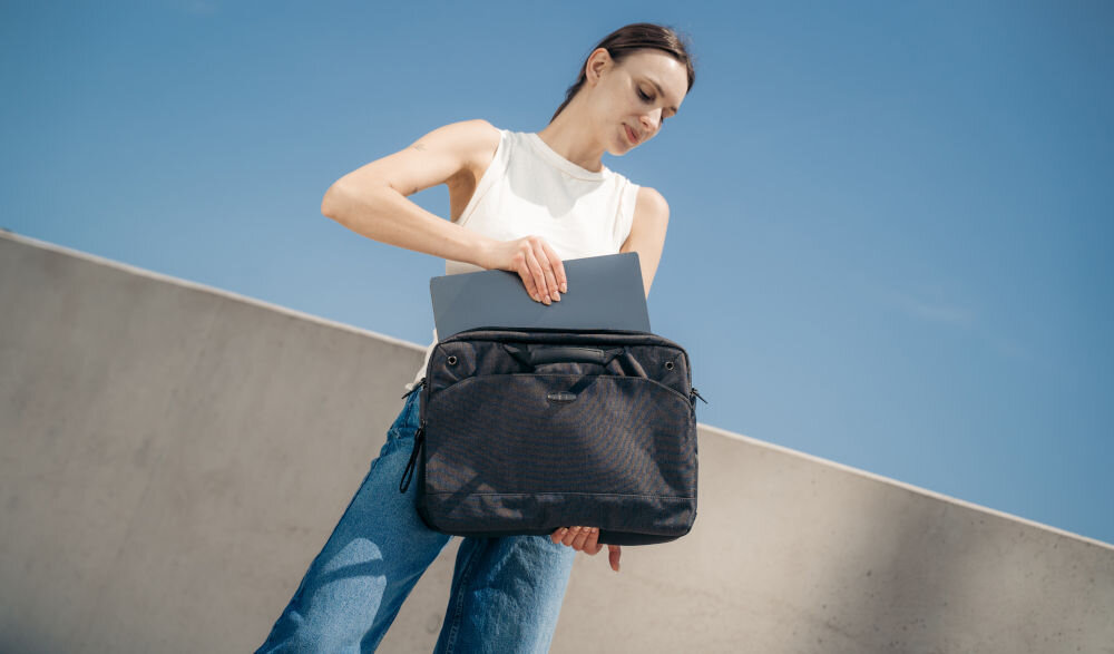 Torba na laptopa ART NB-0362 15.6 cali prezentacja torby trzymanej przez kobietę wkładającą laptopa do niej w tle betonowa ściana i bezchmurne niebo torba bezpieczna w transporcie ochrona laptopa komfort solidne wykonanie transport podróż