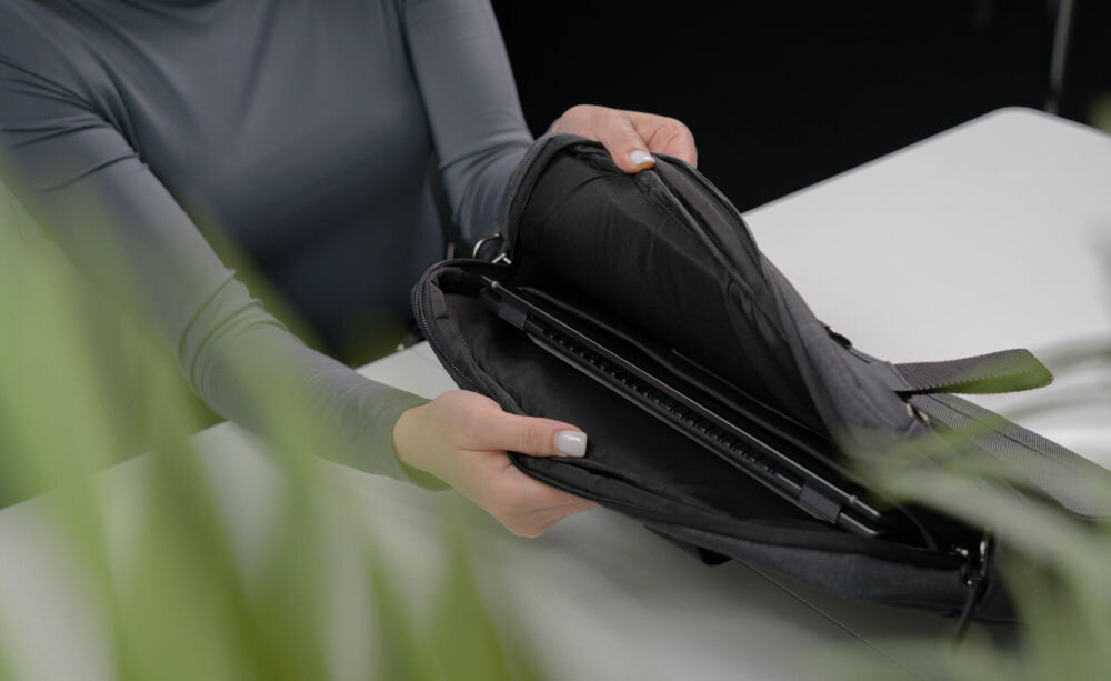 Torba na laptopa ART NB-0362 15.6 cali prezentacja otwartej torby trzymanej przez kobietę w środku laptop w tle białe biurko na przodzie rozmyta roślina miękkie wnętrze chroniące laptop