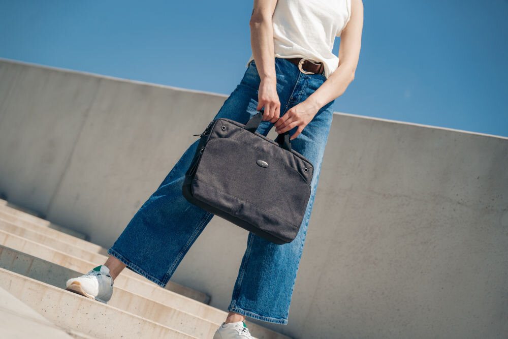 Torba na laptopa ART NB-0362 15.6 cali prezentacja torby trzymanej w ręce za uchwyt przez kobietę stojącej na schodach w tle betonowa ściana i bezchmurne niebo elegancki uniwersalny wygląd praktyczny estetyczny dodatek