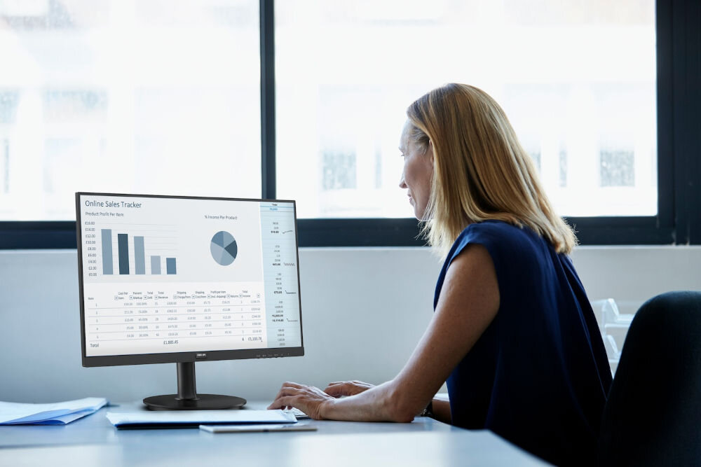 Monitor PHILIPS 241V8LAB 23.8 cala 1920x1080px 100Hz 1 ms [MPRT] kobieta siedząca przy monitorze przy biurku w biurze na ekranie wykresy