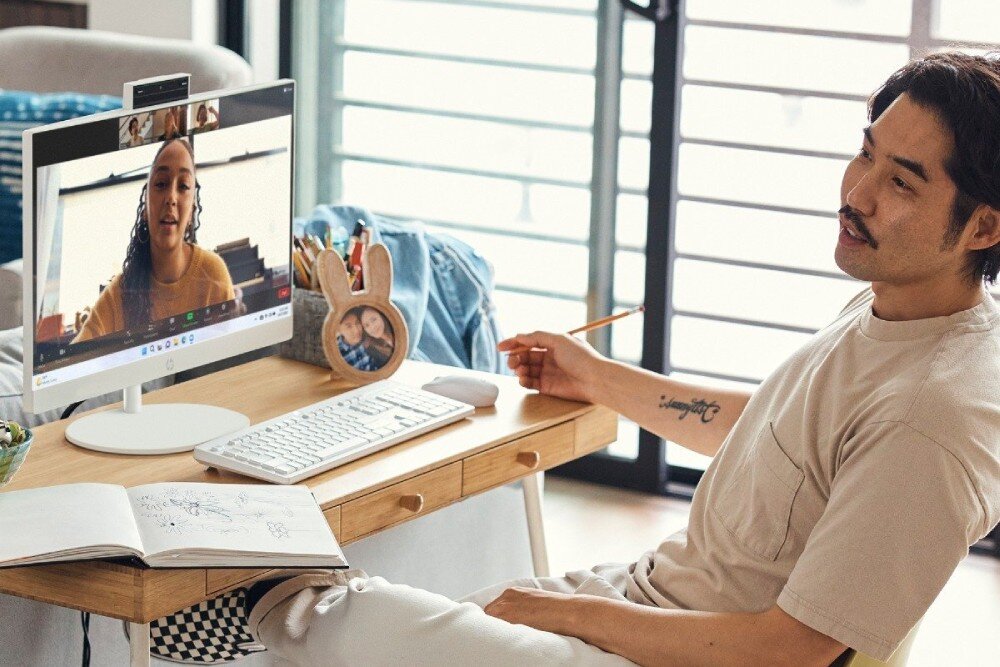 Komputer HP ProOne 240 G10 Mężczyzna siedzi przy biurku podczas wideorozmowy, na ekranie komputera widać kobietę rozmawiającą online. Na biurku znajduje się klawiatura, mysz, notes i ozdobna ramka ze zdjęciem, opis
