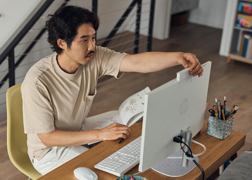 Komputer HP ProOne 240 G10 Mężczyzna reguluje kamerę wbudowaną w tył monitora komputera typu all-in-one. Na biurku obok klawiatury leży otwarta książka i pojemnik z długopisami, podzespoły, procesor, pamięć