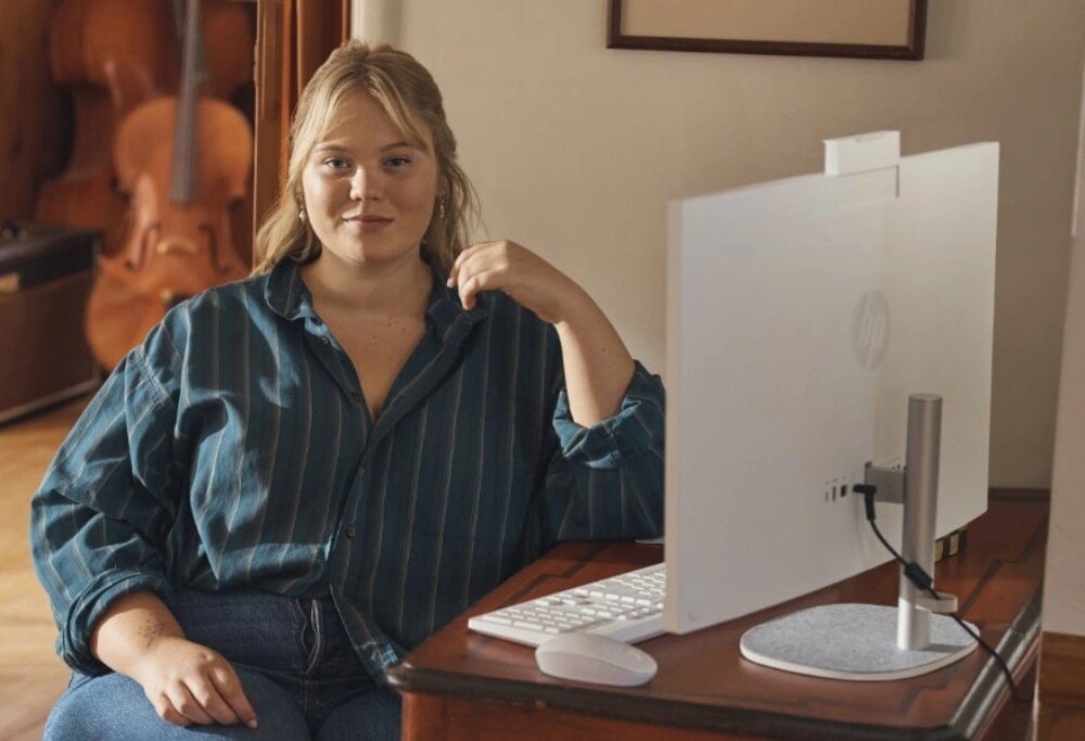 Komputer HP ProOne 240 G10 Kobieta siedzi przy biurku obok komputera typu all-in-one, patrzy w stronę obiektywu z lekkim uśmiechem. Na biurku znajduje się klawiatura i mysz, głośniki, kamera internetowa