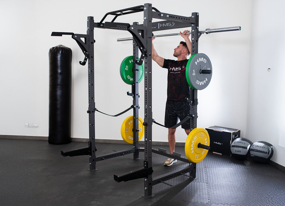 Klatka treningowa HMS KLT3110 pomieszczenie mężczyzna podchodzi do klatki i bierze do ręki gryf po bokach klatki ukazane uchwyty uniwersalność trening w domu