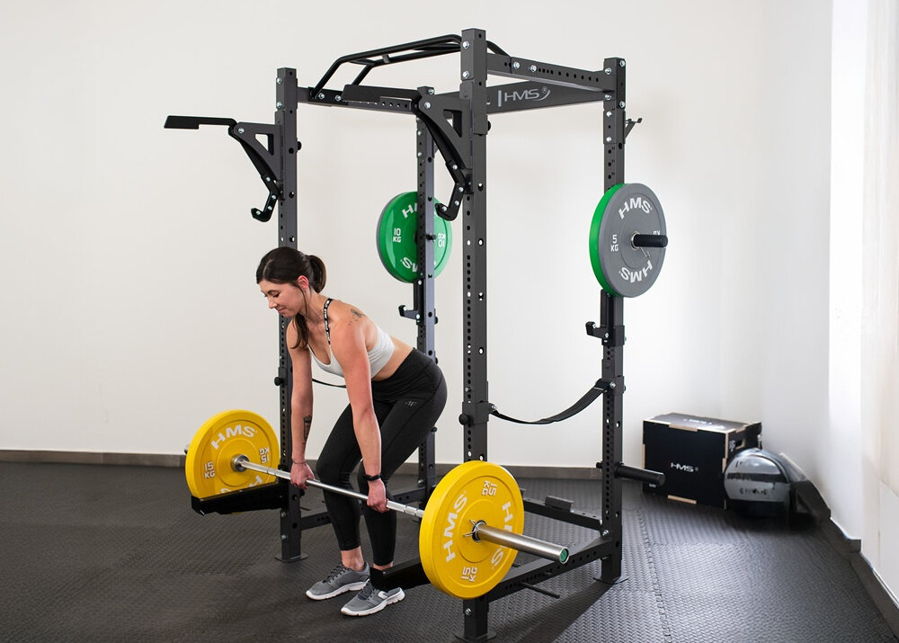 Klatka treningowa HMS KLT3110 pomieszczenie dziewczyna stoi przed klatką i podnosi sztangę, uchwyty amortyzują ewentualne upuszczenie ciężaru profesjonalna strefa ćwiczeń do użytku w klubach fitness i strefach treningu funkcjonalnego stabilna konstrukcja bezpieczeństwo