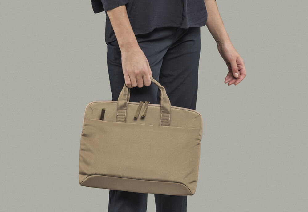 Osoba trzyma beżową torbę na laptopa z dwoma uchwytami. Torba ma prosty, elegancki design i jest wykonana z gładkiego materiału.