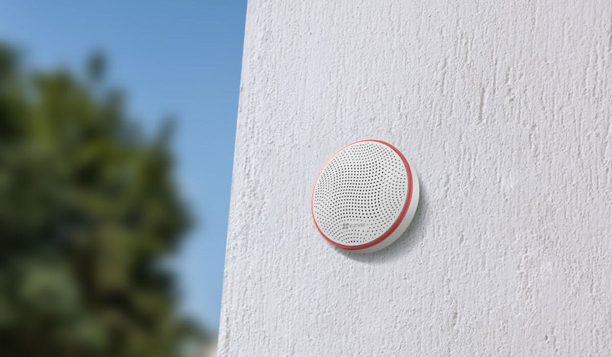 Syrena zewnętrzna EZVIZ CS-T9C-A0-BG, Syrena alarmowa, ochrona domu, łatwa integracja, ustawienia dźwięku, Syrena T9C na ścianie elewacji zewnętrznej, rozmyte zielone tło i błękitne niebo