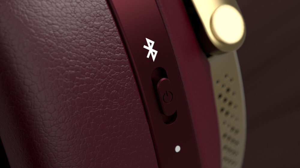 Zbliżenie na bordowe słuchawki z widocznym przyciskiem zasilania i symbolem Bluetooth. Elementy wykończenia są w kolorze złotym.
