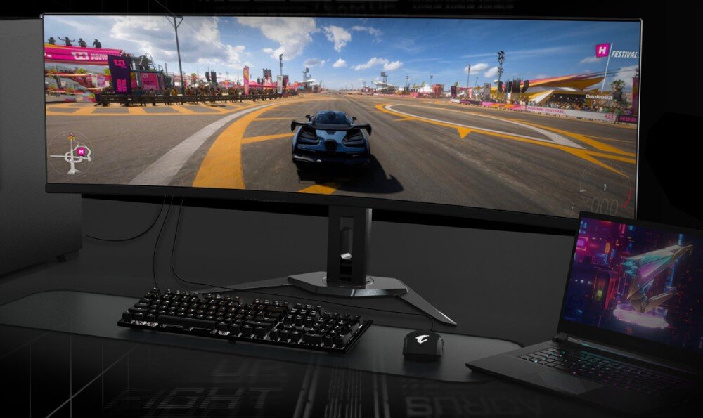 Monitor GIGABYTE AORUS CO49DQ Zestaw komputerowy z dużym monitorem ultraszerokim, na ekranie gra wyścigowa. Obok znajduje się laptop z podłączonym monitorem. Klawiatura i mysz ustawione są na czarnym biurku, przełącznik KVM, obsługa