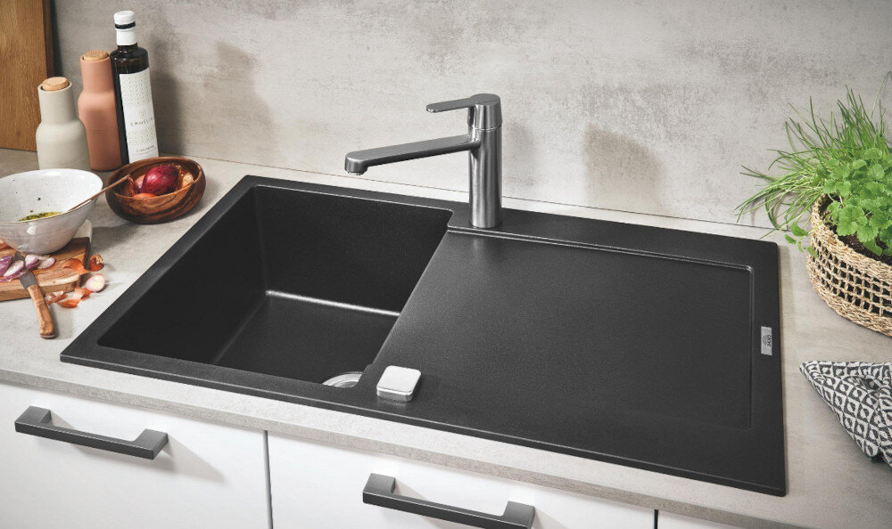 Zlewozmywak jednokomorowy z ociekaczem GROHE K500 31644AP0 Czarny jednokomorowy zlewozmywak wpuszczany w blat z wysoką, chromowaną baterią kuchenną. Wokół stoją produkty spożywcze i dodatki kuchenne tworzące estetyczną aranżację