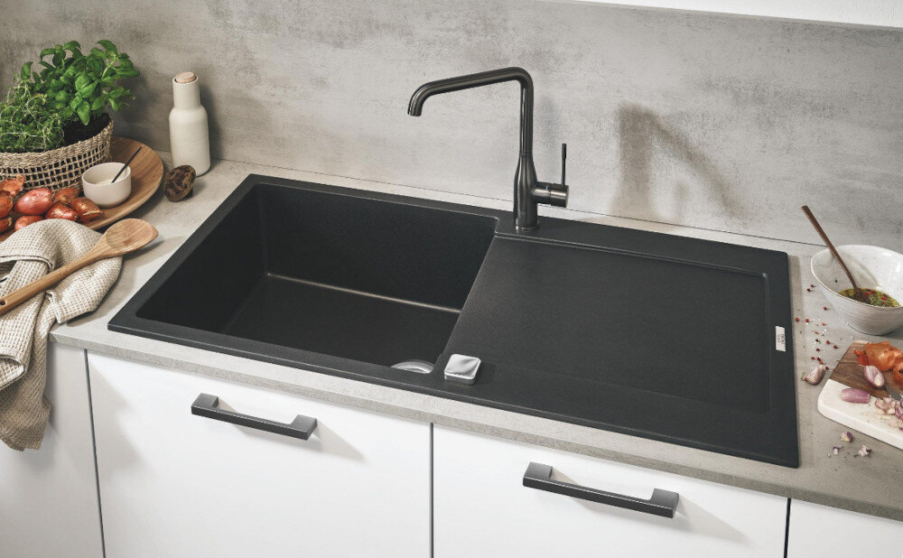 Zlewozmywak jednokomorowy z ociekaczem GROHE K500 31645AP0 Czarny jednokomorowy zlewozmywak wpuszczany w blat z wysoką, chromowaną baterią kuchenną. Wokół stoją produkty spożywcze i dodatki kuchenne tworzące estetyczną aranżację