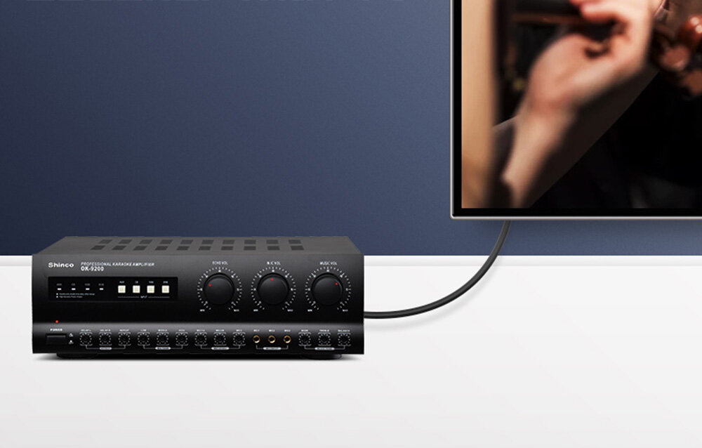 Na obrazie widoczny jest wzmacniacz z napisem \'Shinco Digital Karaoke Amplifier GX-2100\' oraz ekran z fragmentem dłoni trzymającej mikrofon. Wzmacniacz posiada kilka pokręteł i przycisków na panelu przednim.