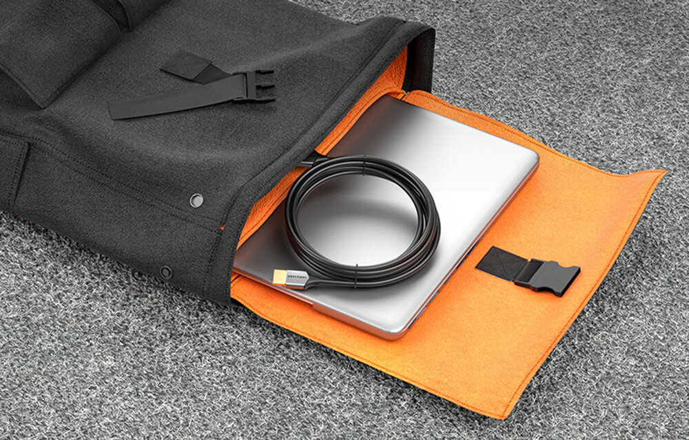 Na obrazie widać czarną torbę z pomarańczowym wnętrzem, w której znajduje się srebrny laptop oraz zwinięty czarny kabel HDMI. Torba leży na szarej powierzchni.