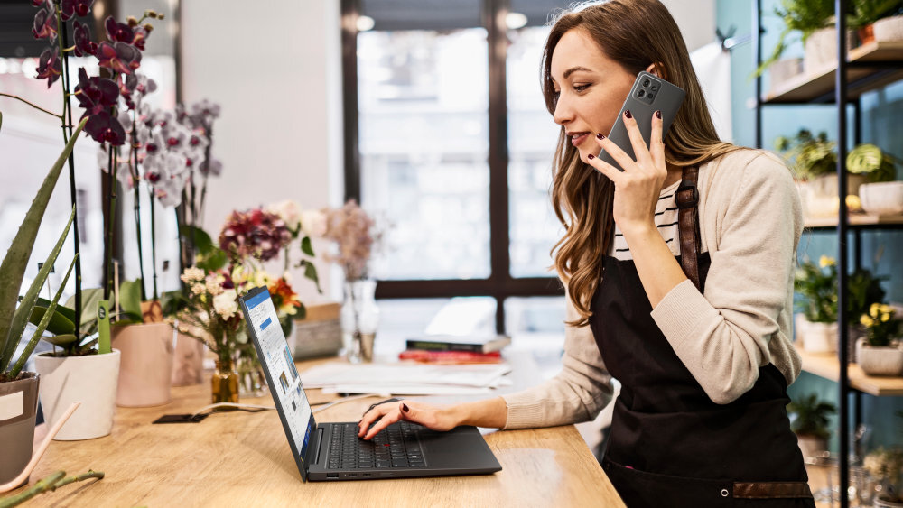 Kobieta w pasiastej bluzce pracuje w kwiaciarni, rozmawiając przez telefon i korzystając z czarnego laptopa Lenovo ThinkPad X1; na ekranie otwarty program biurowy, wokół niej liczne kwiaty i rośliny doniczkowe na półkach. - Wi-Fi 6E (802.11ax) szybkie i stabilne połączenie Bluetooth 5.3