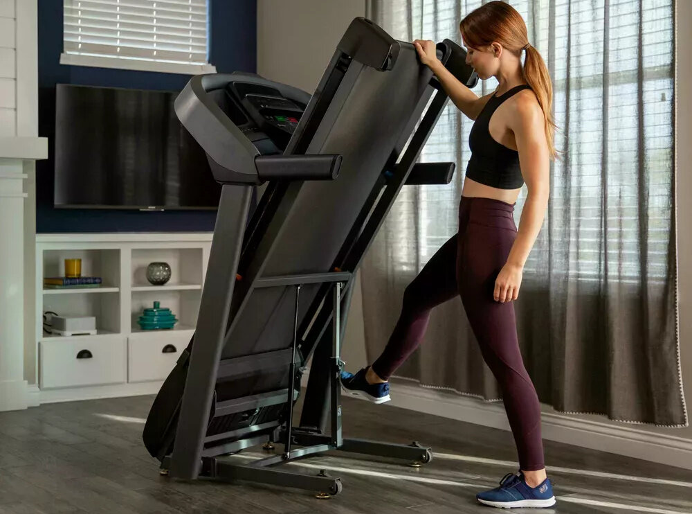 Bieżnia elektryczna HORIZON FITNESS T101 -27 1840W 18km-h pomieszczenie pokój osoba rozkłada bieżnię w pokoju w tle okna telewizor trening w domu wszechstronne zastosowanie kondycja zdrowie