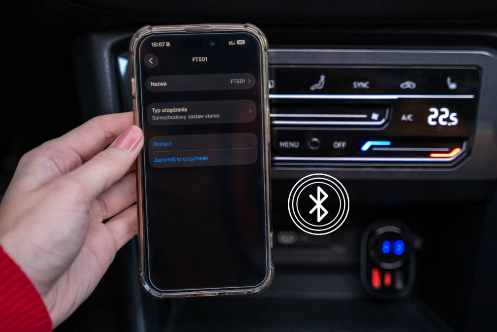 Transmiter FM GOLDENLINE FT501 Dłoń trzyma smartfon, który jest w trakcie parowania Bluetooth z urządzeniem o nazwie FT501. Transmiter jest podłączony do gniazda w samochodzie, pod panelem klimatyzacji.