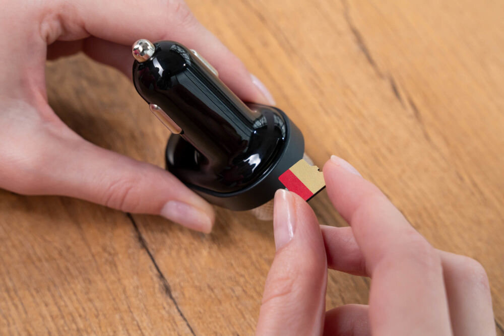 Transmiter FM GOLDENLINE FT501 Dłonie trzymają transmiter FM i wkładają kartę pamięci microSD do jego gniazda, leżąc na drewnianym blacie.