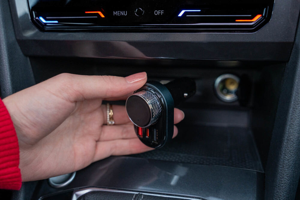 Transmiter FM GOLDENLINE FT501 Dłoń montuje transmiter FM GOLDENLINE FT501 w gnieździe zapalniczki samochodowej, pod panelem sterowania klimatyzacją. Widoczne jest duże pokrętło na transmiterze.