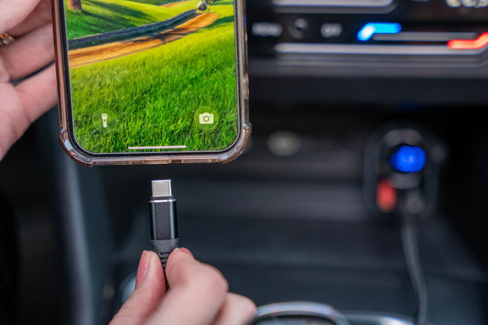 Transmiter FM GOLDENLINE FT501 Zbliżenie na dłoń trzymającą smartfon z kablem USB-C, gotowym do podłączenia do transmitera/ładowarki w samochodzie. W tle widoczny jest podłączony transmiter.