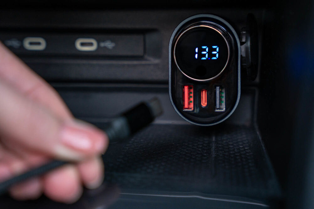 Transmiter FM GOLDENLINE FT501 Dłoń zbliża kabel USB-C do portu USB w samochodzie. Transmiter FM podłączony do gniazda zapalniczki samochodowej wyświetla na cyfrowym ekranie napięcie akumulatora: 13.3. Widoczne są dwa czerwone i jeden zielony port USB w transmiterze.