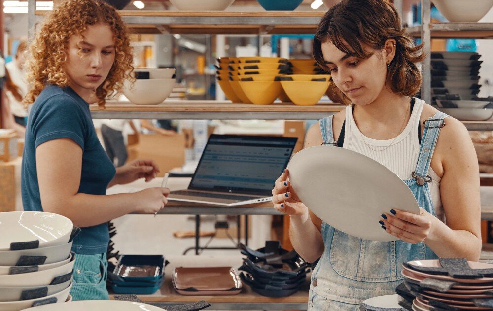 Laptop HP ProBook 440 G10 Dwie kobiety w sklepie z ceramiką – jedna trzyma talerz i dokładnie go ogląda, druga w tle patrzy na ekran laptopa, który stoi na półce z naczyniami, opis