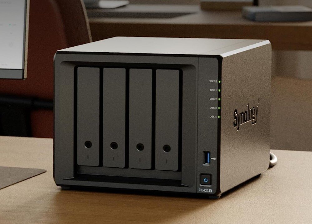 Serwer plików SYNOLOGY DiskStation DS423+ Czarny serwer plików o czterech zatokach na dyski stoi na drewnianym biurku. Na panelu przednim znajdują się diody LED sygnalizujące status urządzenia. W tle widać monitor i elementy biura, opis