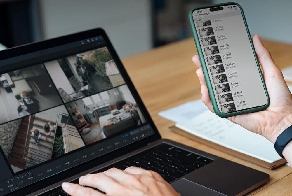 Serwer plików SYNOLOGY DiskStation DS423+ Osoba trzymająca smartfon z otwartą aplikacją do monitoringu, na której wyświetlone są nagrania z kamer. W tle ekran laptopa z podglądem na żywo z kilku kamer, synchronizacja, aplikacja, wersjonowanie, porty LAN