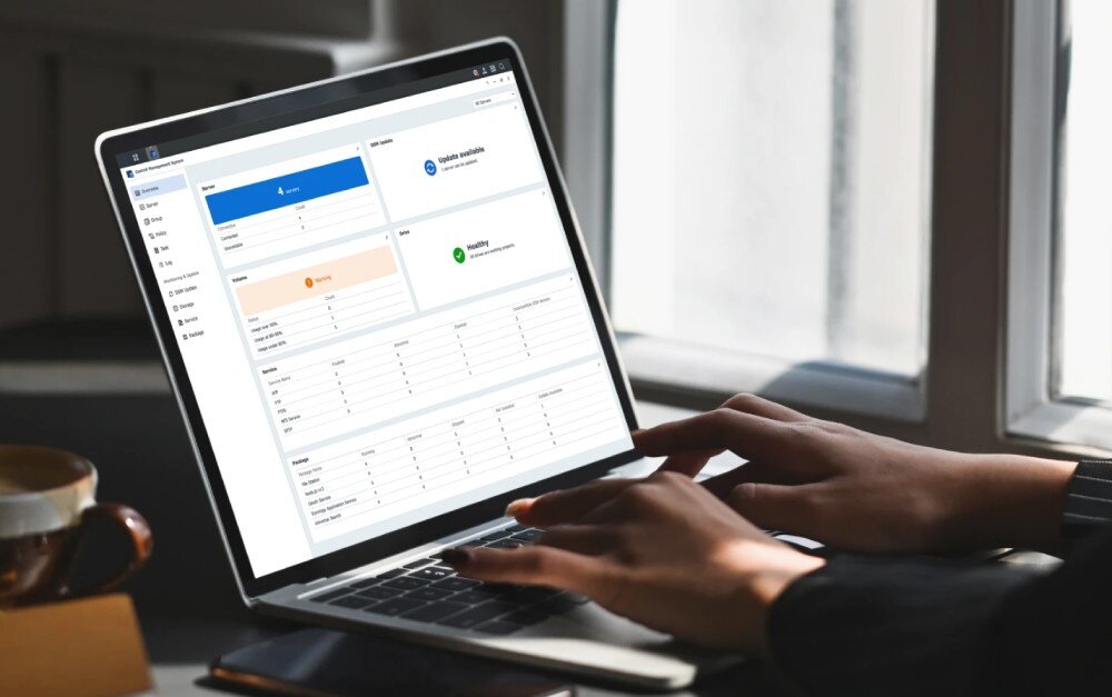 Serwer plików SYNOLOGY DiskStation DS423+ Osoba korzystająca z laptopa z otwartym interfejsem zarządzania serwerem plików. Na ekranie widać statystyki systemu, informacje o aktualizacjach oraz status dysków, system operacyjny, zarządzanie, tryby RAID